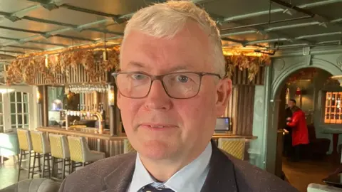 Michael Lennon who has white hair and brown glasses looks into the camera as he stands in front of a hotel bar. He is wearing a light blue shirt, tie and brown jacket.