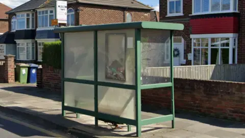 Green bus shelter with rubbish piled up in a black frame on window which is meant for bus timetable. The side of the shelter is dusty and missing a window pane. There are semi-detached houses behind the shelter.