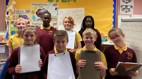 A group of eight children from Victoria Junior School, smiling and holding clipboards with their poems on.