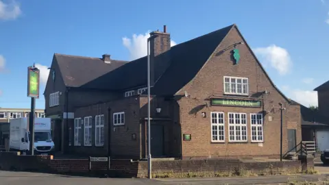 BBC The exterior of the Lincoln Imp pub in Scunthorpe. The dark brick building is bathed in sunshine with blue skies. A van is parked next to a large green sign.
