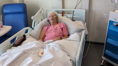 A woman in a pink top with short grey hair lies in a hospital bed.
