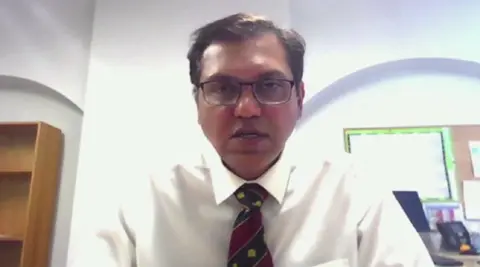 Hemant Nemade with medium-length dark hair and glasses. He is wearing a white shirt and red and blue tie and looking at the camera. There is a bookshelf to the left behind him and a noticeboard to the right.
