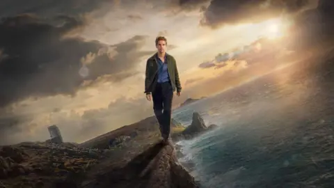 UKTV Damien Molony as Det Jim Bergerac walking along a cliff with the sea to his left and Jersey to his right. The sky is cloudy and the sun is setting behind him. He is wearing a green jacket with a blue shirt and black trousers.