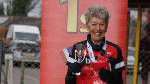 Anne Dockery wearing red, black and white sports gear. She smiles at the camera as she holds a silver trophy in one hand and a red finisher t-shirt in the other. She has a medal with a white ribbon around her neck and is standing in front of a red 1st place banner.