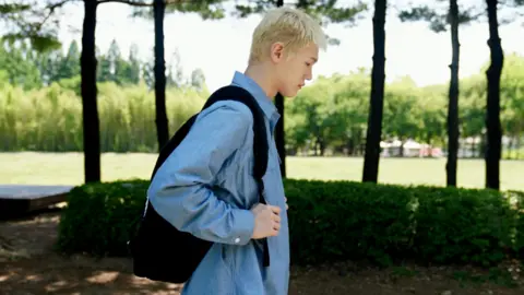 BBC Korean/Jungmin Choi Bain dressed in a long sleeved blue shirt carrying a black backpack, poses for a photo with trees in the background