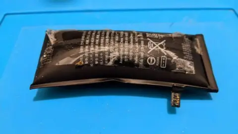 Jamie Farnell / Shropshire Phone Repairs There is a black lithium ion battery on a light blue surface. The battery has swolen, and includes worn out lettering.