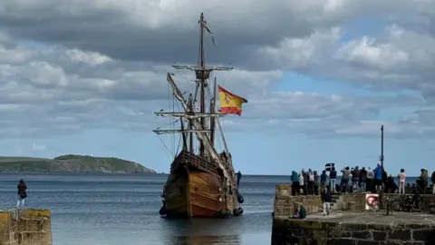 A tall ship with a Spanish flag. It is sailing in the water. The sea is a deep blue. There are people watching it from the harbour. The sky is blue with grey clouds. 