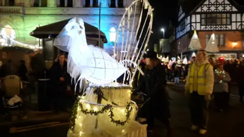 An illuminated dove leads the lantern parade through Chester. The dove is on top of a cake and wearing a veil.