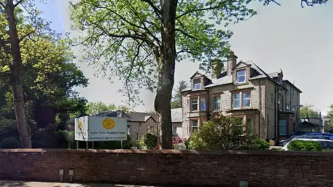 A Google Street View image of the entrance to Ostley House in Barrow - a double-fronted period property, behind a low wall with greenery in the front driveway.