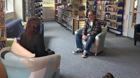 A man sitting across from a girl in a chair. In the background is rows of bookshelves.