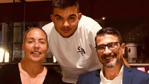 Alex Aghili Kordmahale Alex is sat next to her husband with her son perched behind them. They are sat at a dining table.