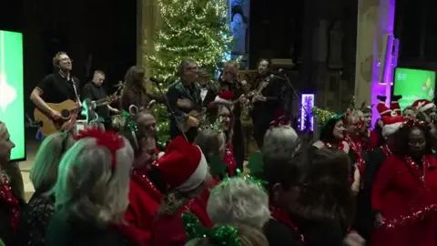 Ed Young/BBC A picture of the Everly Pregnant Brothers on stage in Sheffield cathedral with members of the NHS Voices Choir in front of them wearing red and white hats and tinsel.