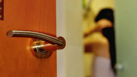 A woman in a white towel stands with her back to an open door. The door is seen in focus, while the figure of the woman is blurred. She has long dark hair.