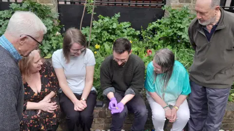 Epping Forest District Museum CIO Six people looking at an Anglo-Saxon gold ring, which is held in the gloved hands of the man in the middle. Two men are standing to either side of the group. Four people - two women, a man and then another woman - are seated on a low wall. Behind them is a flower bed. 