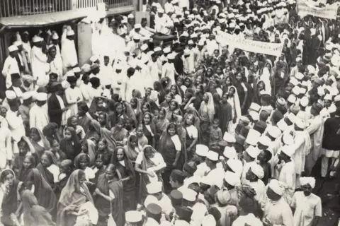 The Alkazi Collection of Photography A procession led by women to encourage hand spinning in Mumbai passes through a busy street in India. The women are all wearing saris and have their heads covered. They are surrounded by men, most of whom are wearing caps and kurtas, on both sides of the street. 