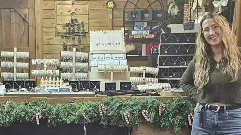 Alex Healy A woman with long blonde wavy hair stands by a festive market stall displaying jewellery smiling. 