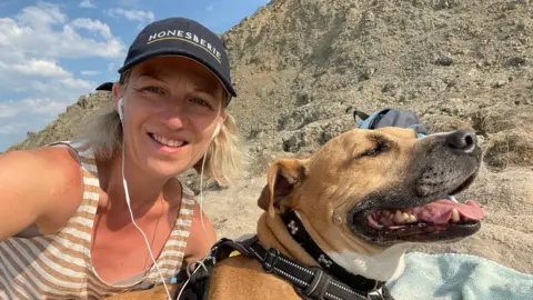 Laura Bayliss Bayliss is smiling in next to her dog at Holywell Bay. Bayliss is wearing a black cap, headphones and a striped vest. 