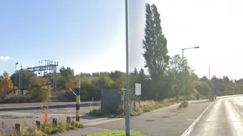 A street view of a road with a concrete site to the left and a railway line in the distance.
