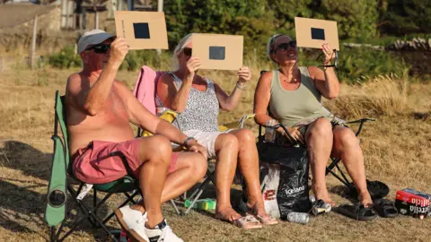 Three people sitting on camping chairs hold what looks like pieces of black glass in frames of cardboard to the sky to watch the partial eclipse