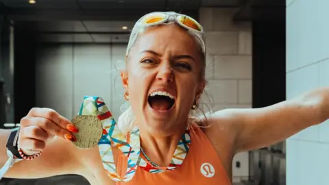 Anya Culling is wearing an orange running vest with her blonde hair tied up and white sunglasses on her forehead. She is holding a Tokyo Marathon 2026 medal in her right hand, with a colourful lanyard around her neck and her left arm in the air in celebration.