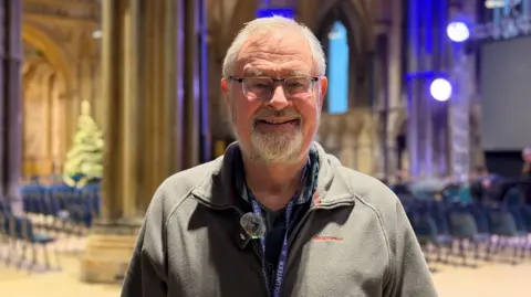 Amber Gash/ BBC Brian Porter is standing in Lincoln Cathedral and smiles at the camera. He has grey hair, a beard and moustache. He is wearing glasses and a grey half-zipped fleece. He has a lanyard around his neck. 