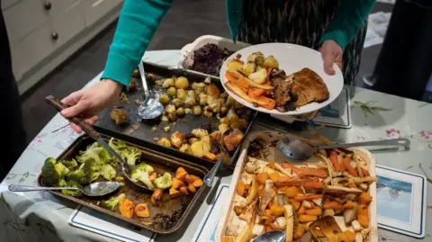 A close up of one hand holding a plate of food containing a Christmas dinner of chicken, potatoes and vegetables, the other hand with a serving spoon scooping vegetables off a tray on a table which also contains two other trays of vegetables.