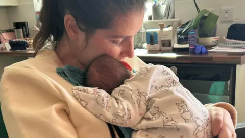 Lucy Lintott Smith Lucy Lintott Smith holds to her chest sleeping baby SS, who is wearing a Pooh onesie, in their kitchen.