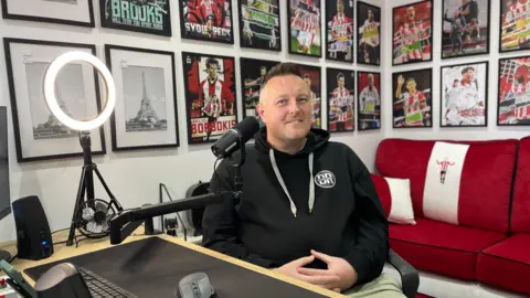 Mark Ansell/BBC A man with short hair and wearing a dark hoodie sat in a recording studio. A mic is close to his face. Behind him is a red sofa and posters of footballers on the walls, as well as some pictures of the Eiffel Tower.
