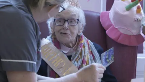 Brian Johnson An elderly man in a birthday sash is shown a card by a nurse.