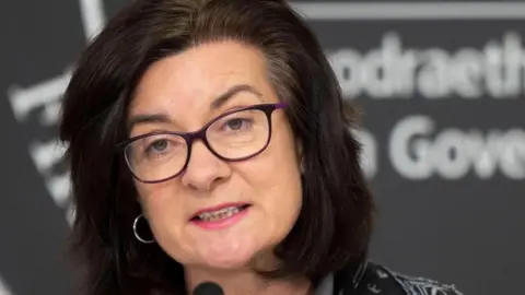 A close-up of Eluned Morgan at a Welsh government news conference. The white lettering of the Welsh government logo can be partly seen in blurred form in Welsh and English behind her.