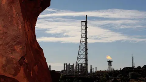 Getty Images Uma plataforma de rocha vermelha em primeiro plano com um local industrial em segundo plano, incluindo uma torre de gás flamejante