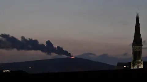 Pacemaker Smoke rises and drifts from a crest of flames on a mountain. A church steeple is in the middle ground. 