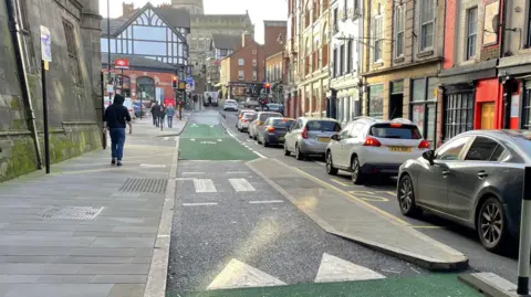 A green cycle lane with a pedestrian crossing in the middle leading to a narrow bus stop. Cars can be seen stuck in traffic in a lane on the right-hand side of the bus stop. Pedestrians are walking on the pathway to the left of the image.
