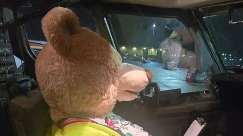 Nikki Gaynor Poppy is a large beige-furred teddy bear wearing a high visibility vest and looking at a clipboard while sitting in the cockpit of an aeroplane.