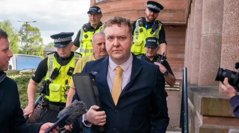 PA Media Jordan Linden outside court. He has short brown hair and is wearing a dark blue suit, light pink shirt and a yellow tie, and carrying a black folder. There are a number of police officers behind him and reporters to the side.