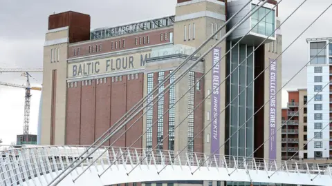 The Baltic Centre For Contemporary Art is a tall red-brick building on the River Tyne. Near the top, black bricks are laid in way that spells Baltic Flour Mills. The white Millennium Bridge is in the foreground of the photograph.