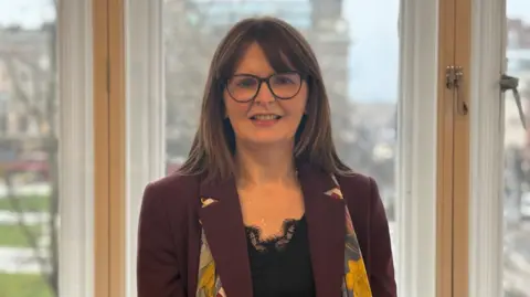 Caoimhe Archibald is standing in front of large windows and is smiling at the camera. She has shoulder-length hair and is wearing a pair of large-framed glasses. She is also wearing a burgundy coat, a black top and a floral patterned scarf.