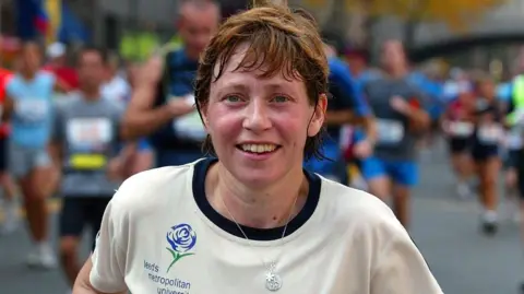 Chris Ison/PA Jane Tomlinson running along a road with many other runners behind her. She is wearing a cream-coloured top marked "Leeds Metropolitan University" and is wearing a necklace with a star. She smiles at the camera.