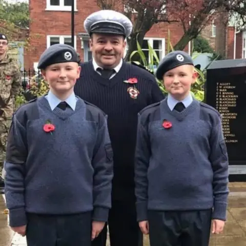Karl Shannon A man with two boys, his sons. The man is wearing a navy blue lifeboatman uniform and white naval-style cap. The boys are wearing cadet uniforms, with blue jumpers, light blue shirts, dark blue ties and black berets. All three have poppies pinned to their chests. A war memorial be seen, with a red-brick house in the background.