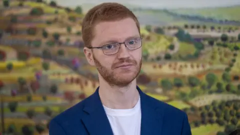 PA Media Ross Greer, who has short ginger hair and glasses, in front of an animated picture of green fields. 