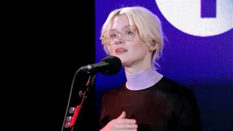 Maisie Peters sings in the Live Lounge. She has blonde hair and gold glasses 