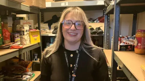 A blonde woman standing in a small storage room lined with metal shelving units. On the shelves around them are a variety of groceries. The woman is wearing a dark top and a lanyard with various badges or keys attached as well as some square glasses