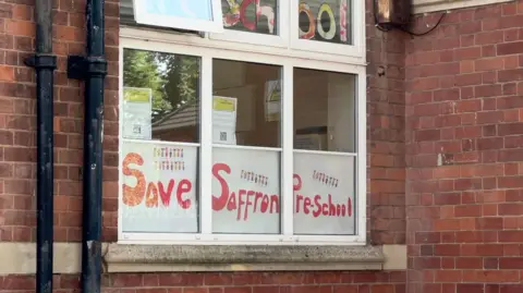 The photo shows a window with the words Save Saffron Pre-school in red lettering.