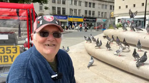 A man smiling into the camera with a red cap on and pigeons in the background.