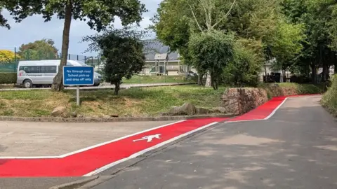 In the image there is a bold red-painted pathway section marked by a white pedestrian symbol and surrounded by white lines. There is a school building and playground in the background. There is a small lane to the right with lots of trees. 