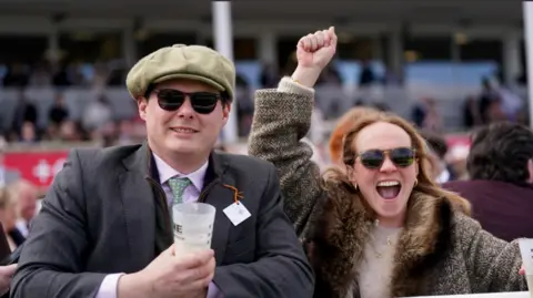 PA Media A man, holding an empty beer glass, smiles as a woman beside him celebrates at winner at Aintree