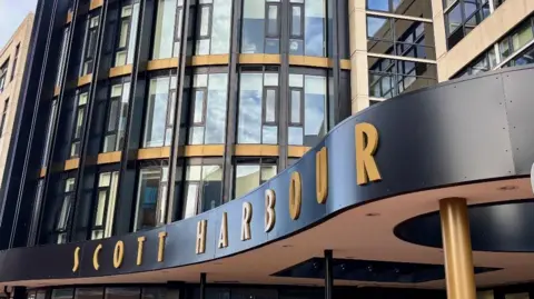 Cardiff Council the exterior of a modern building with a curved black canopy featuring large gold letters spelling “SCOTT HARBOUR.” The building façade is composed of tall glass windows framed by black vertical beams and accented with gold horizontal bands, creating a sleek and contemporary design. The structure rises several stories high, with a mix of glass and beige stone elements. Above the canopy, the windows reflect a partly cloudy sky, adding depth to the scene. A single bird is visible in the sky near the top of the building, and the overall lighting suggests a bright day with natural sunlight.