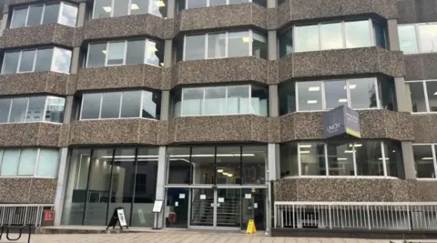 Front aspect of a five storey building, with brown pebble dash walls. The front entrance is in shot at the middle of the picture. The pavement in the foreground.