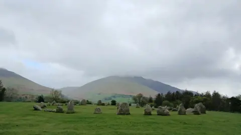 Jackdog A circle of grey, mossy stones arranged on a grassy field. There are green mountains in the distance and a large grey cloud over head. It is so low, the top of the mountain would be misty.