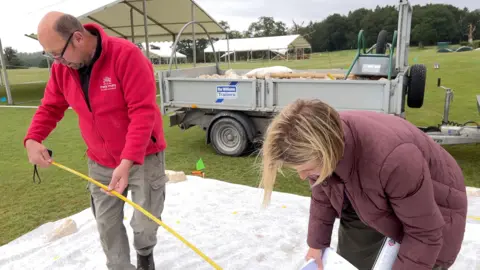 A man in a red fleece and woman wearing a padded coat, use a tape measure to transfer the garden plan onto the site, in front of a trailer filled with material for the build.